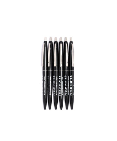 6 black plastic ballpoint pens with metal clips and retractor buttons. Field Notes logo in white.